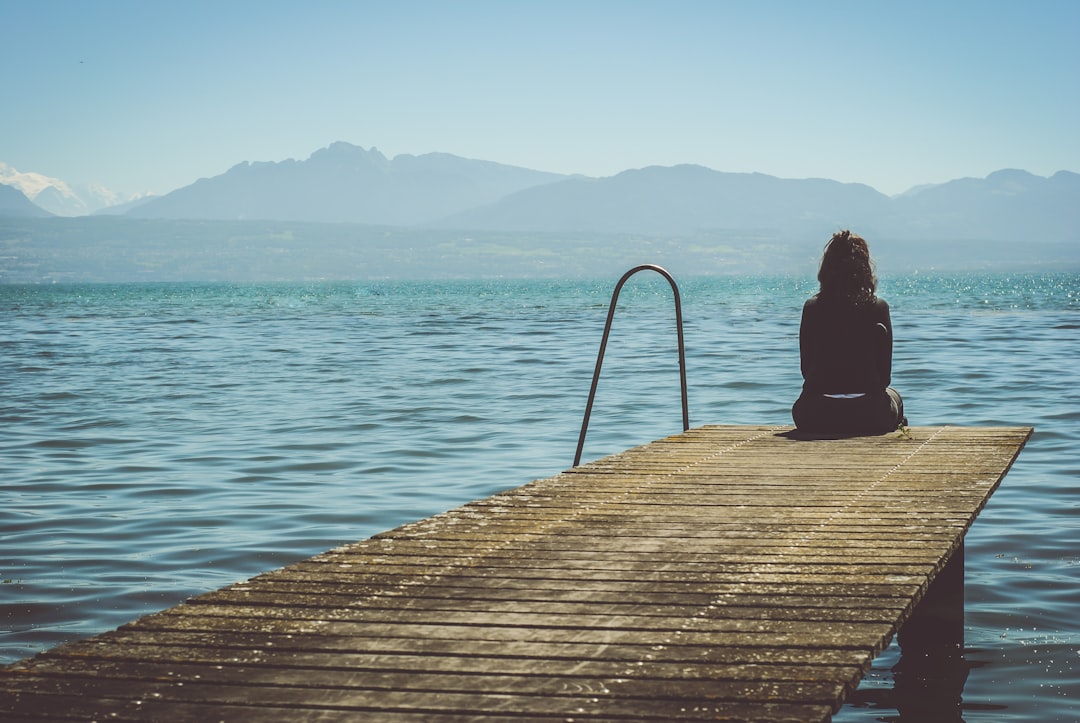 A person sits peacefully on the edge of a wooden dock in the early morning light, looking out over calm water, representing the contemplative choice of a waterfront lifestyle.