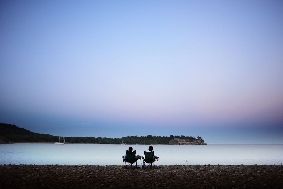 A couple sits in comfortable chairs on the grassy bank of their property, peacefully enjoying the view of the river in front of their home.